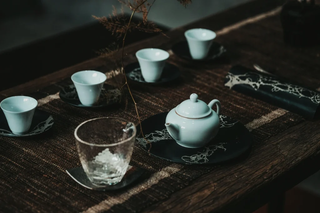 A serene tea setting on a rustic wooden table featuring a white teapot, five small cups on dark saucers, and a clear glass. Elegant, calm ambiance.