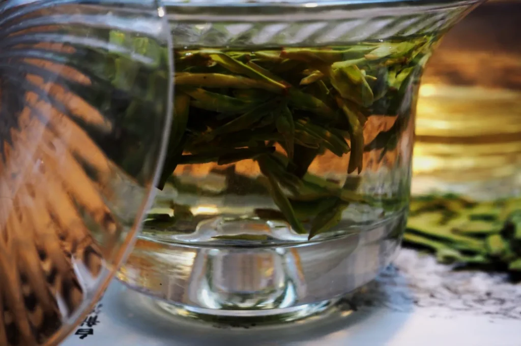 A glass teacup with a lid, filled with steeping green tea leaves. The setting is serene and warm, suggesting a tranquil tea-drinking moment.