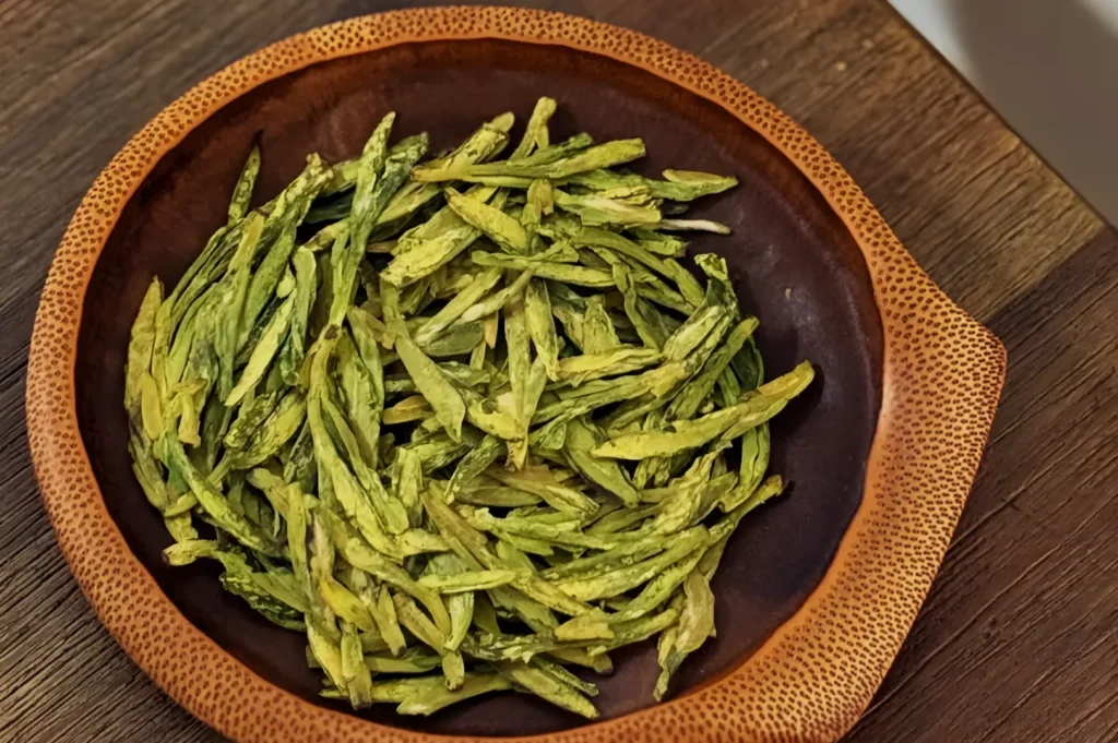 A brown ceramic bowl filled with long, dried green tea leaves sits on a wooden table. The vibrant green contrasts with the bowl's rustic appearance.