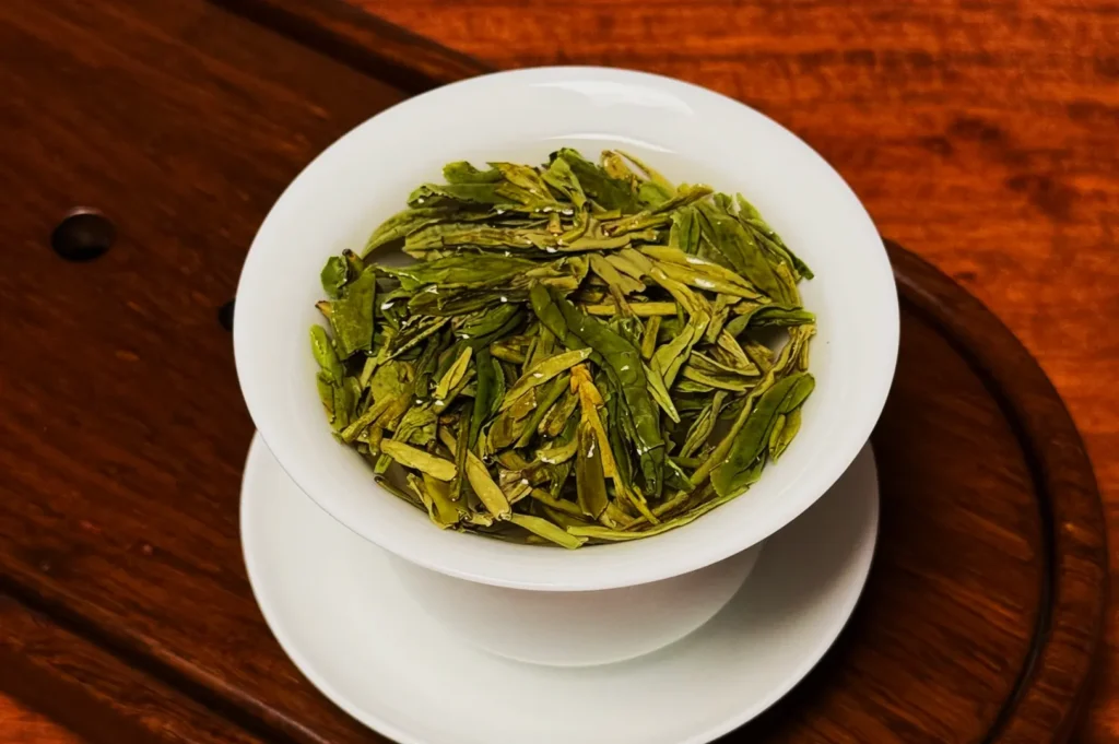A close-up of a white teacup filled with vibrant green tea leaves placed on a saucer with a warm wooden table background, conveying freshness and tranquility.