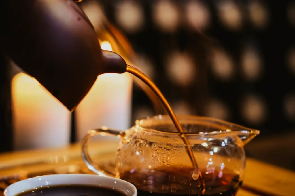 A clay teapot pours tea into a glass teapot, with steam rising. In the background, soft candlelight creates a warm, cozy atmosphere.