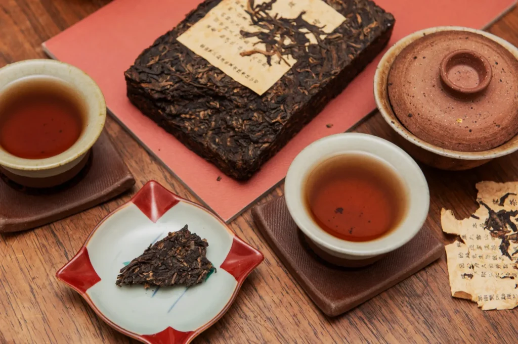 Two cups of dark tea sit on wooden coasters beside a clay teapot. A block of pressed tea and a torn paper with characters add an authentic, rustic feel.
