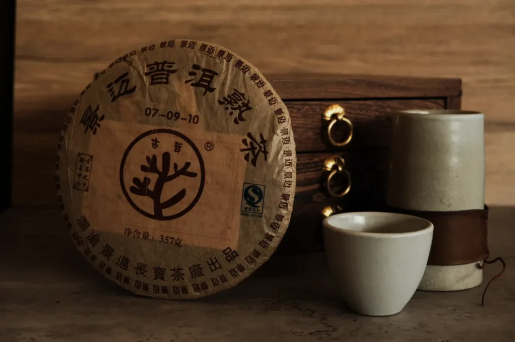 A round, wrapped pu-erh tea cake with Chinese characters rests against a wooden chest with gold handles. Nearby are a ceramic cup and a leather-bound container, creating a traditional, rustic tea setting.