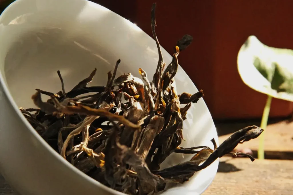 Loose dried tea leaves spill from a white ceramic bowl, with soft sunlight illuminating their textures. A blurred green leaf is visible in the background.