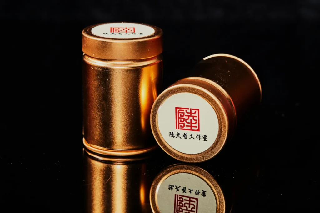 Two cylindrical bronze canisters with white labels and red Chinese characters are displayed on a reflective black surface, suggesting elegance.
