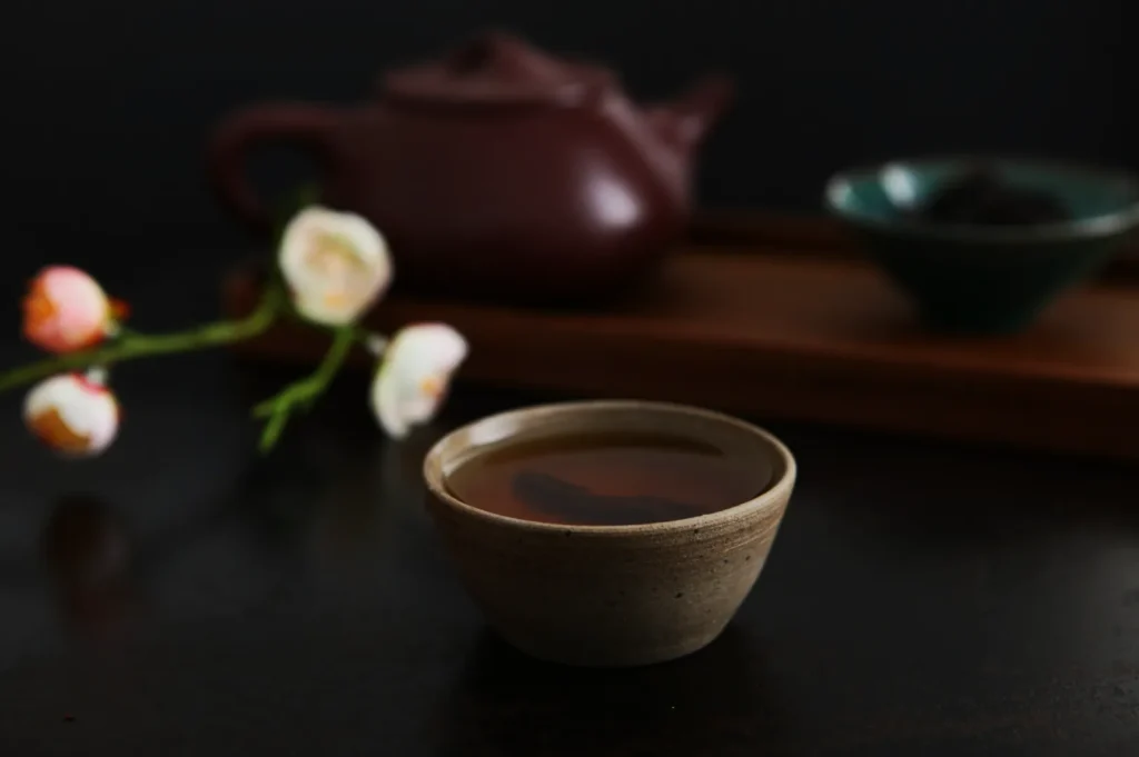 A ceramic teacup filled with tea sits on a dark surface. Behind it, a red teapot, a wooden tray with a green bowl, and soft-focus pink flowers add warmth.