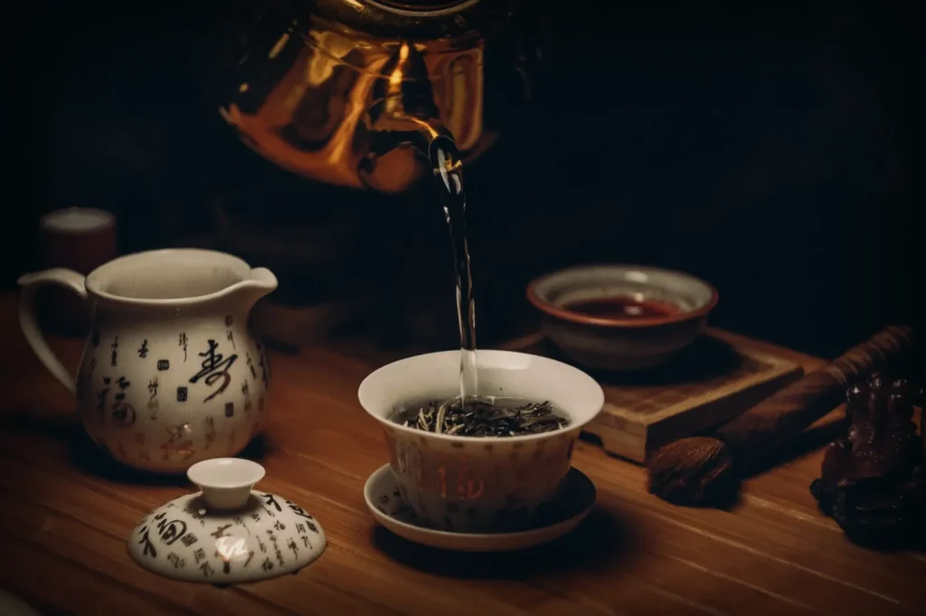 A golden teapot pours hot water into a bowl of tea leaves on a wooden surface. Nearby, a teapot with Chinese characters and a small cup add an authentic touch.