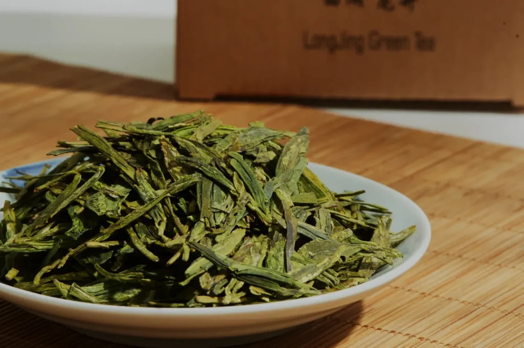 A pile of flat, dried green tea leaves sits on a white plate against a bamboo mat. A cardboard box labeled "Longjing Green Tea" is in the background.