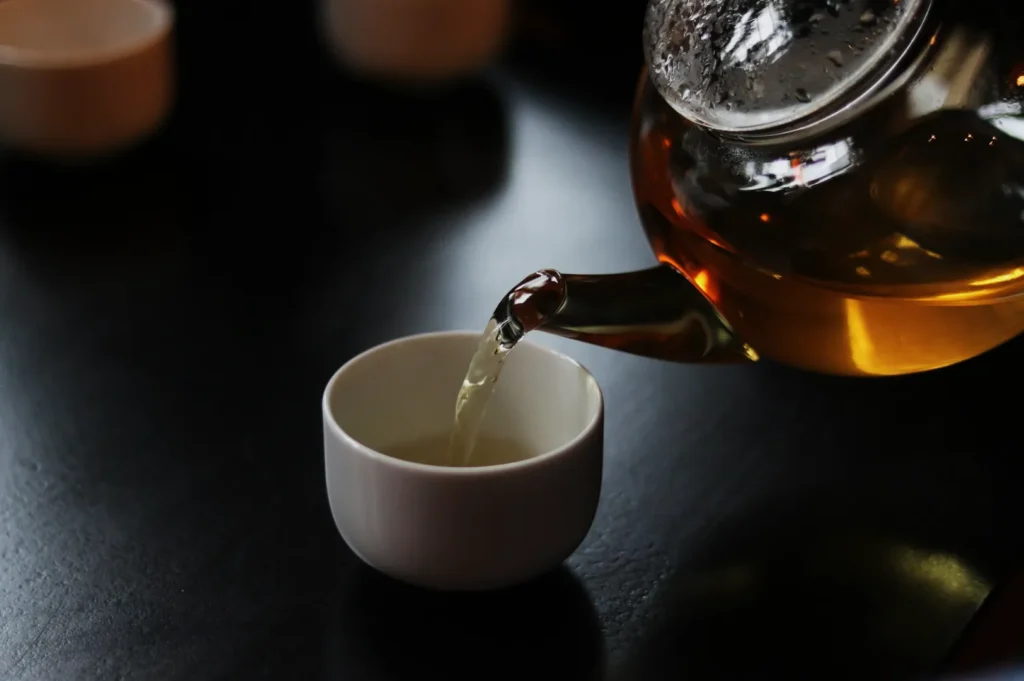 A transparent teapot pours golden tea into a small white cup on a dark table, creating a warm and calming ambiance with soft reflections.
