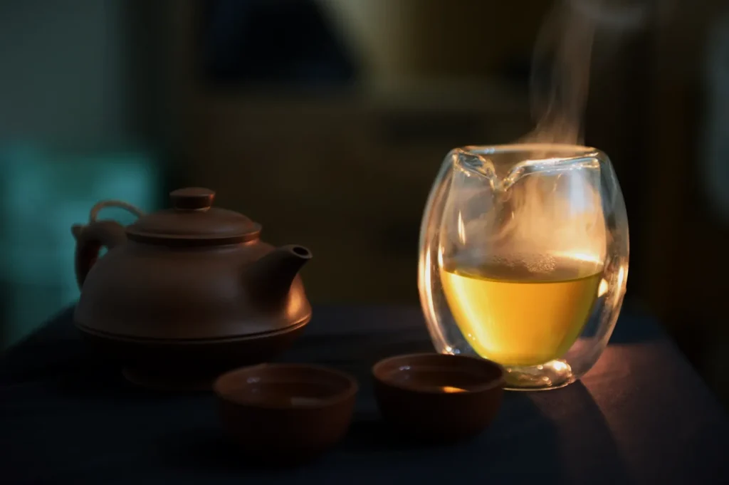 Steaming glass of tea beside a brown clay teapot and two cups on a dark cloth, creating a warm, calm, and inviting atmosphere.