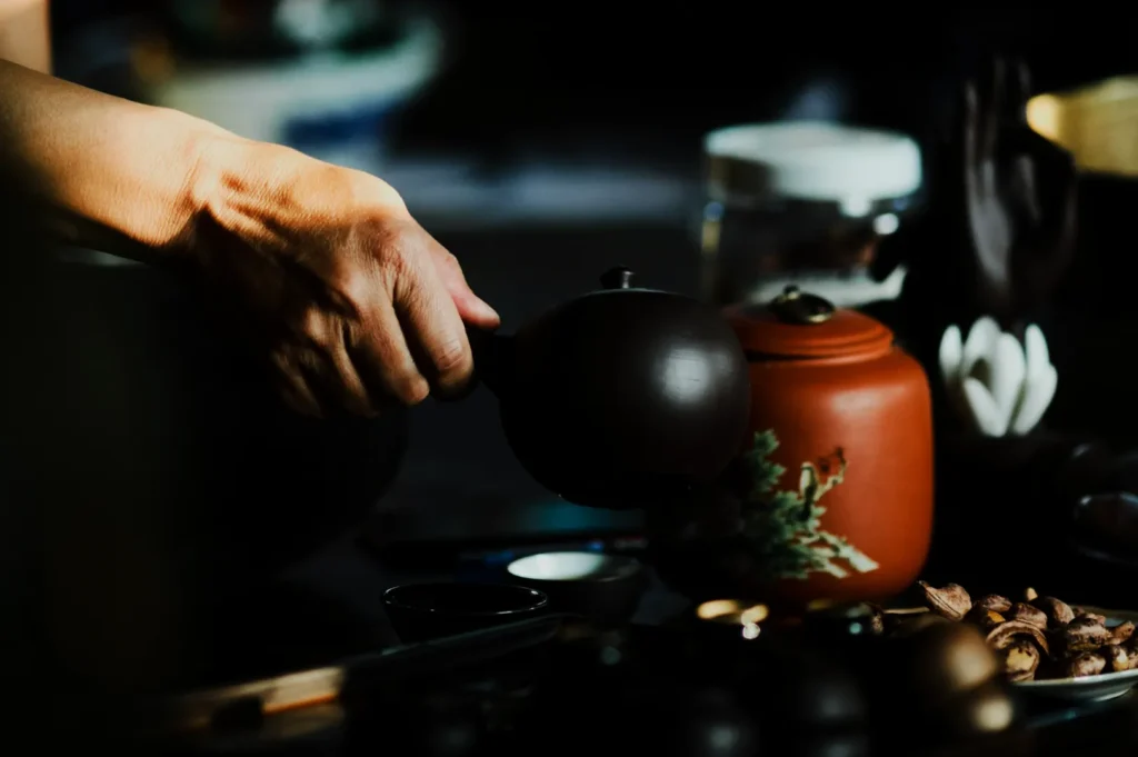 A hand pours tea from a brown teapot into a small cup. Nearby is an ornate orange teapot and scattered nuts, creating a calm, cozy atmosphere.