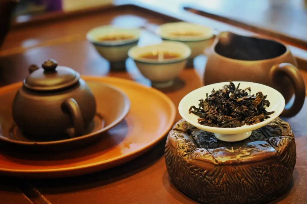 Close-up of a traditional tea set with a clay teapot on a tray, a bowl of loose leaves beside, and steaming cups in the background, creating a serene mood.