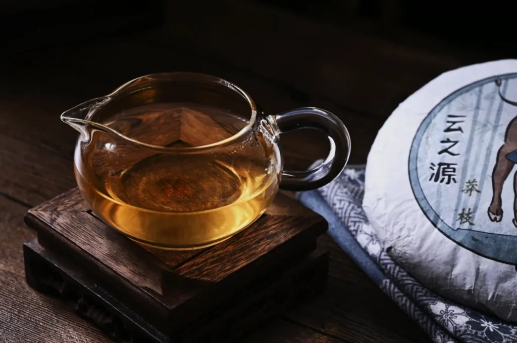 A glass teapot with golden tea sits on a wooden stand. Beside it, a wrapped cake of Pu-erh tea with Chinese characters on a cloth. Warm, cozy ambiance.