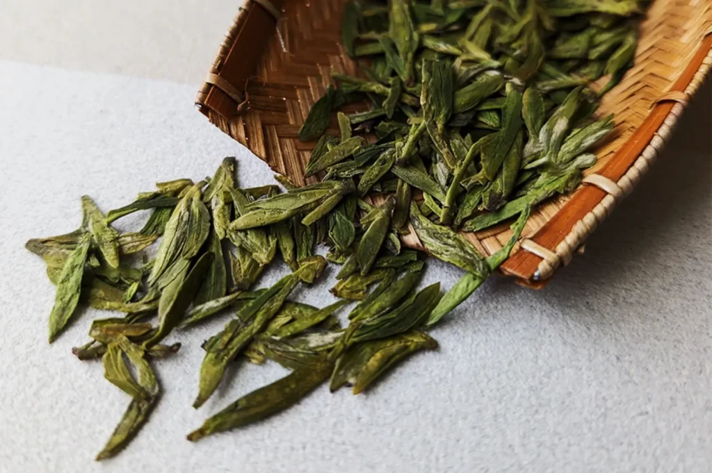 Loose green tea leaves spill from a woven bamboo basket onto a light surface. The dried leaves are elongated and vibrant, suggesting freshness.