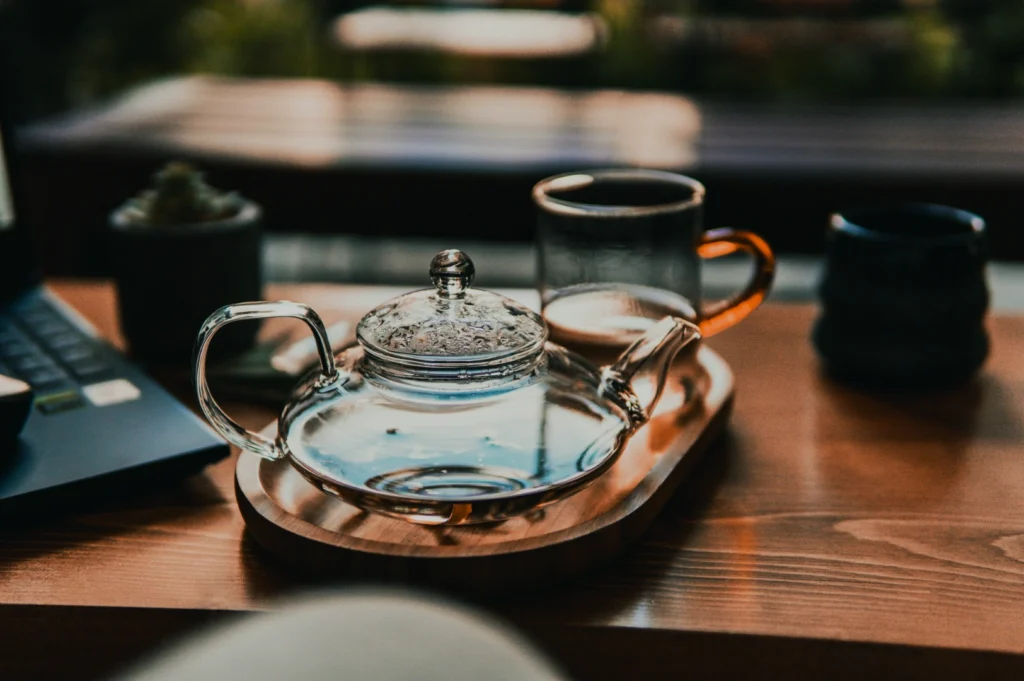 Glass teapot and two mugs on a wooden tray beside a laptop on a table. Soft natural light and a cozy, relaxed atmosphere are conveyed.