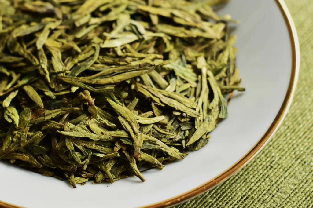 Close-up of dried, green tea leaves on a white plate with a gold rim. The textured, green woven mat underneath adds an earthy feel.