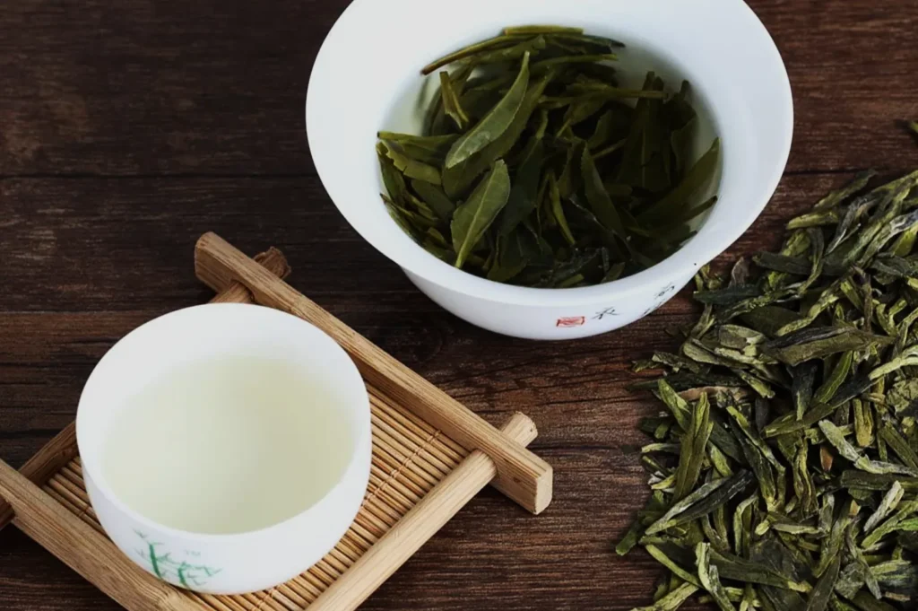 A white cup of pale green tea sits on a bamboo coaster, next to a bowl of brewed tea leaves. Dried leaves are scattered on the wooden table.