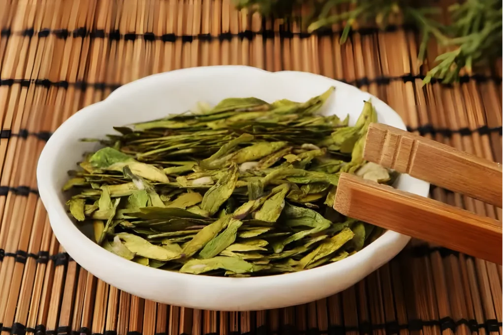 White bowl filled with loose green tea leaves, placed on a bamboo mat. Chopsticks rest on the bowl's edge, conveying a traditional, serene tea setting.