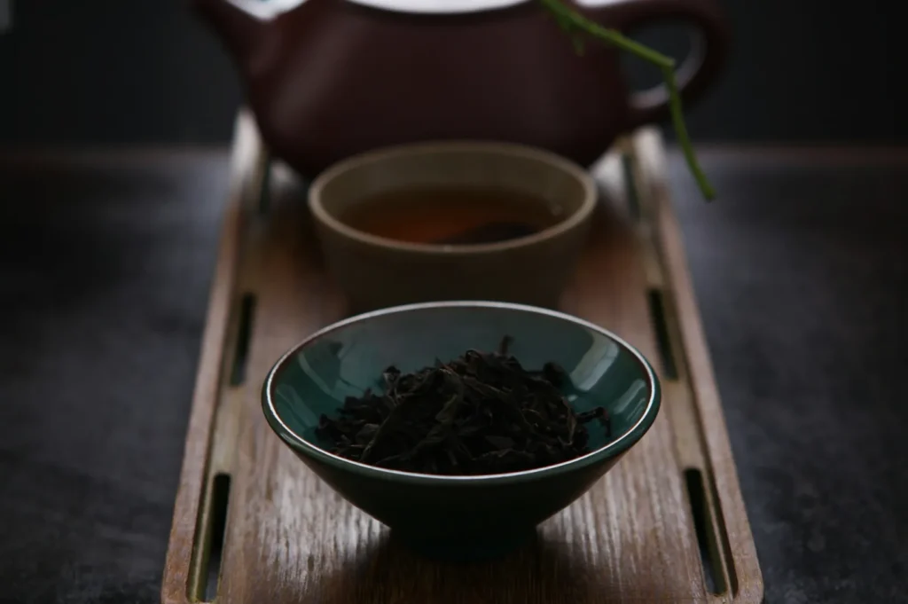 A serene tea setup with a bowl of loose leaf tea in the foreground, a cup of brewed tea, and a clay teapot on a wooden tray, evoking a calm ambiance.