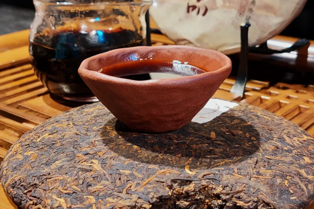 A rustic clay teacup filled with dark tea sits on a large disk of tea leaves. Behind, a glass teapot is partially visible on a wooden tray.