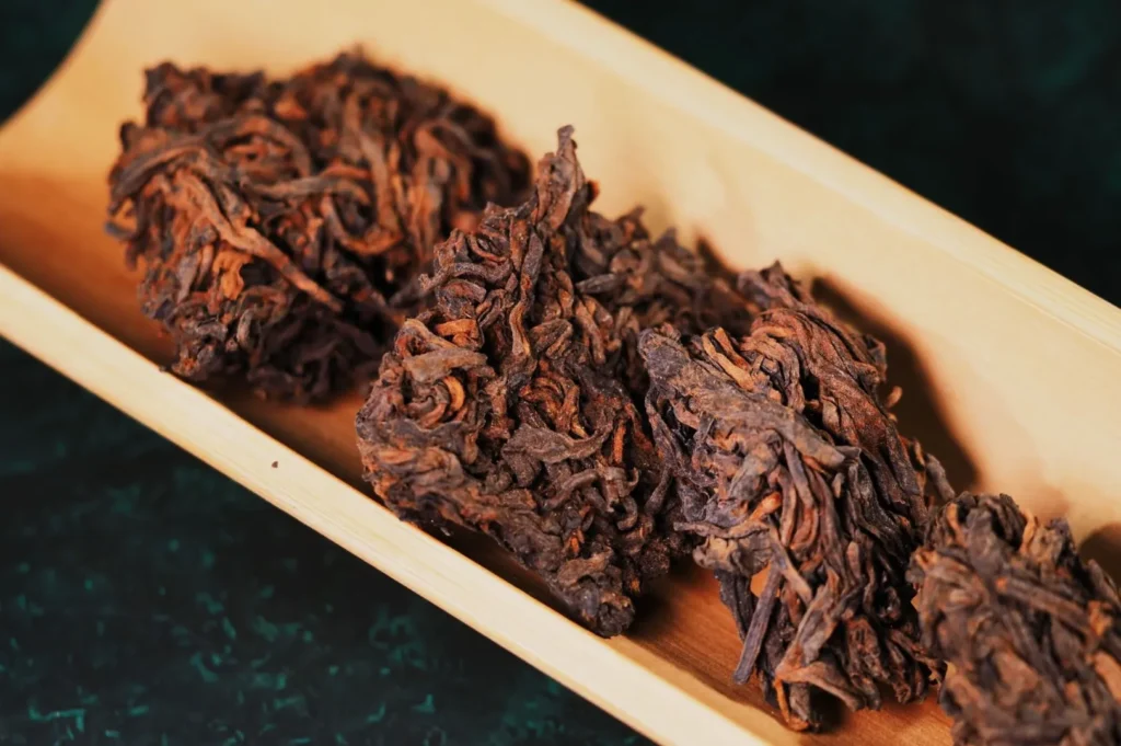 Dried, twisted tea leaves on a light wooden tray placed against a dark green textured surface. The arrangement conveys an earthy, organic feel.