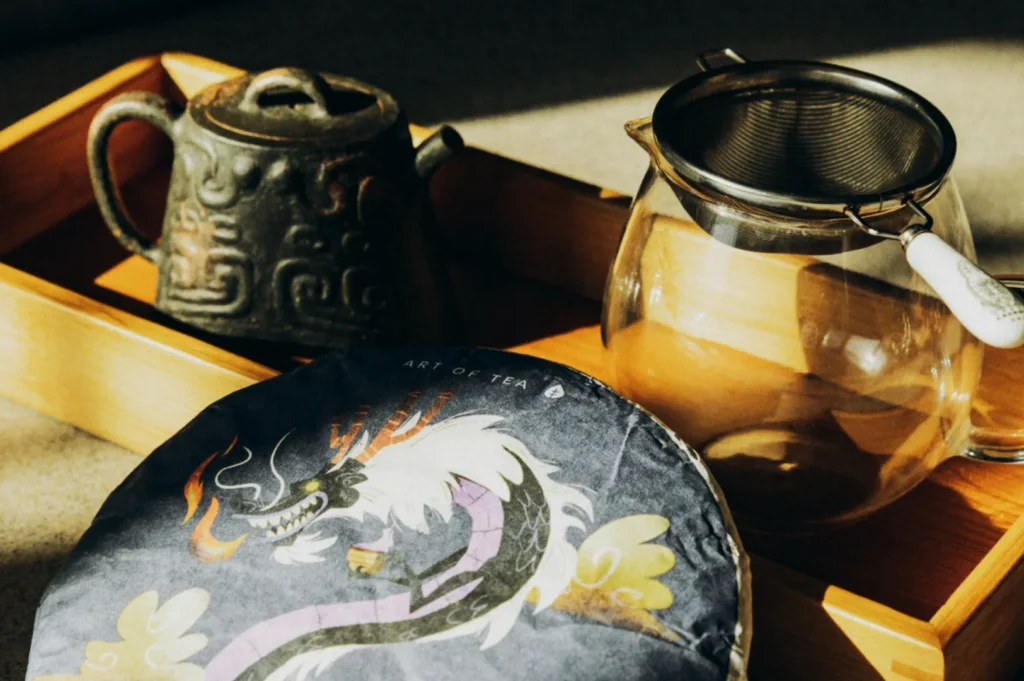 A rustic teapot, clear glass pot, and tea strainer sit on a wooden tray. A tea package with a dragon design adds a traditional and artistic touch.