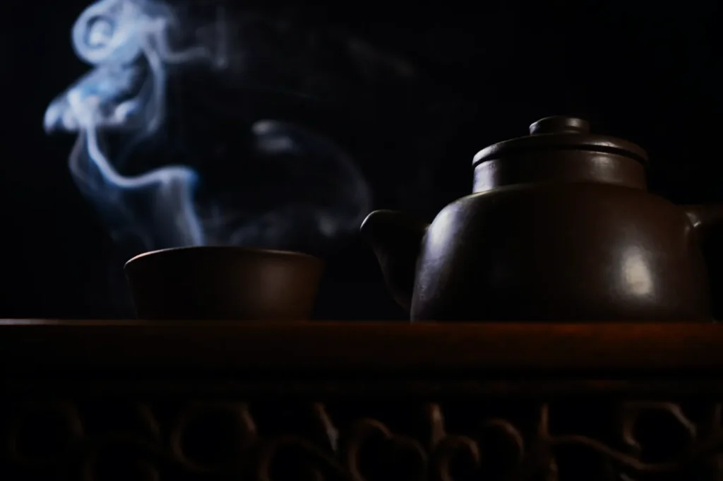A clay teapot and cup on a wooden table, with swirls of steam rising into the dark background, creating a calm and serene atmosphere.