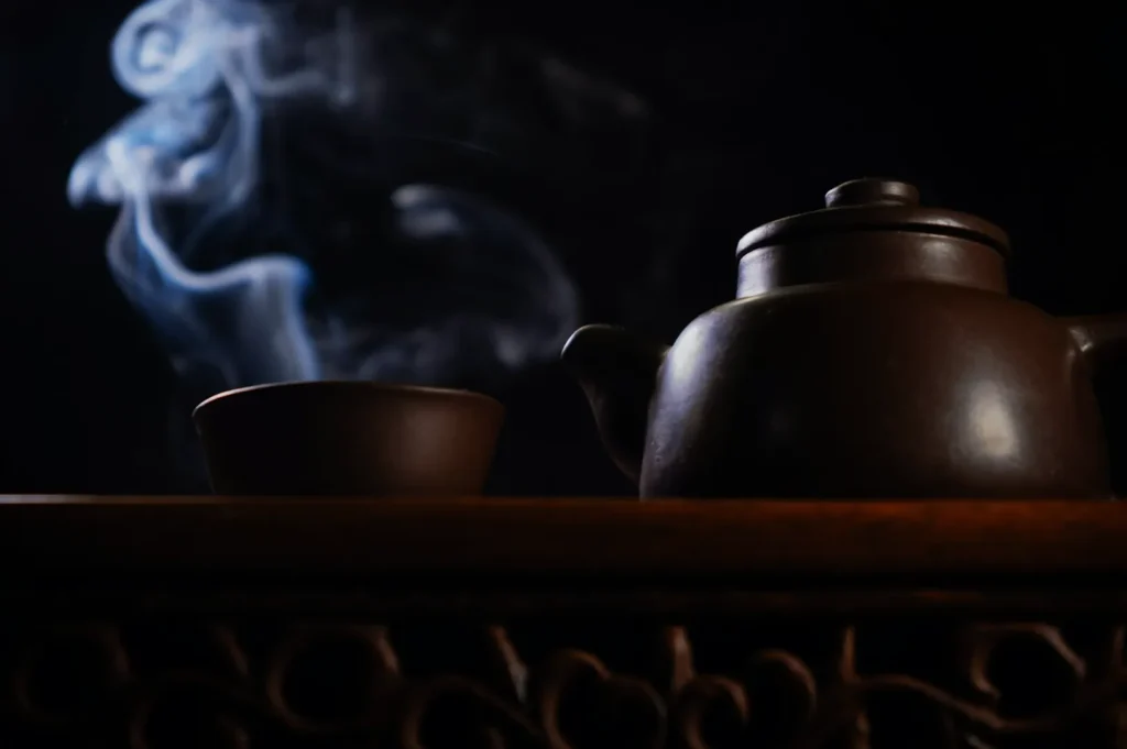 A dark, moody scene with a clay teapot and cup on an ornate wooden table. Wisps of steam rise gracefully against the black background, evoking warmth.
