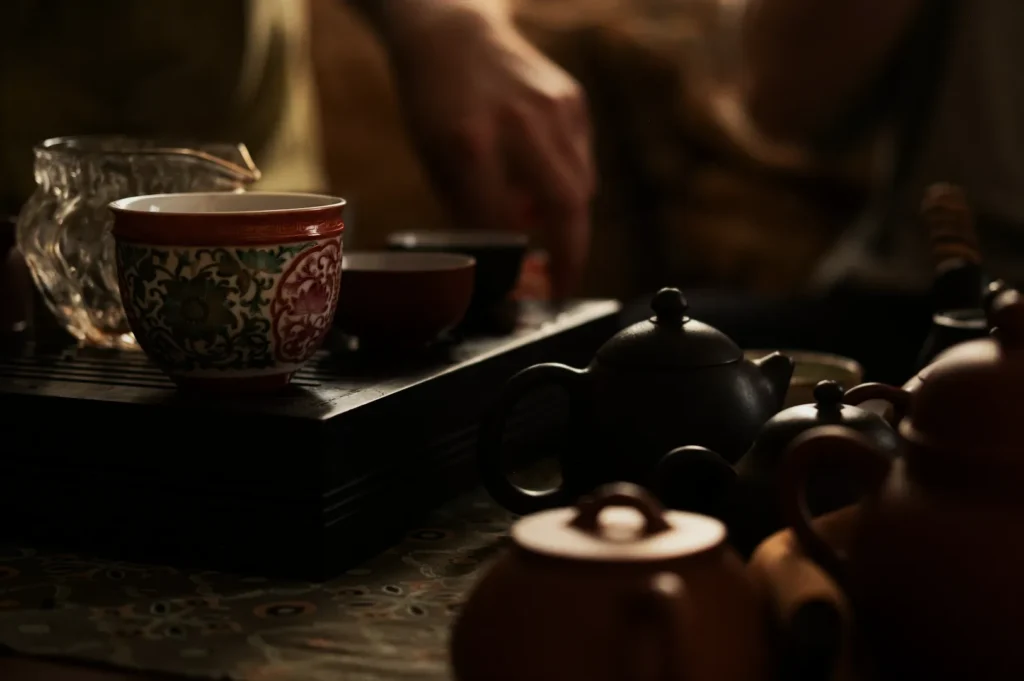 A serene tea set scene with ornate floral cups on a black tray, surrounded by dark teapots. A person's hand gently reaches for a cup, evoking calmness.