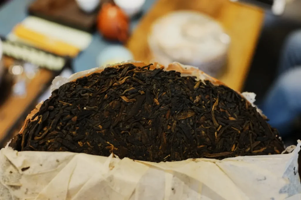Close-up of a round, dark Pu-erh tea cake partially wrapped in white paper. The tea's texture appears dense and leafy, suggesting rich aroma and flavor.