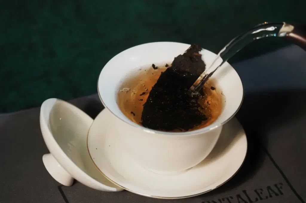 A white cup with a lid holds dark pu-erh tea leaves while hot water is poured in, creating a rich infusion against a dark green background.