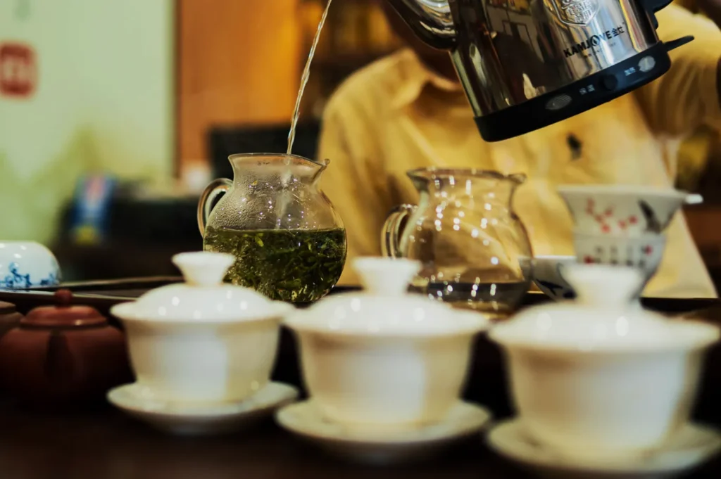A person pours hot water from a kettle into a glass teapot with green tea leaves. Ceramic teacups and pots are in the foreground, suggesting a serene tea ceremony.