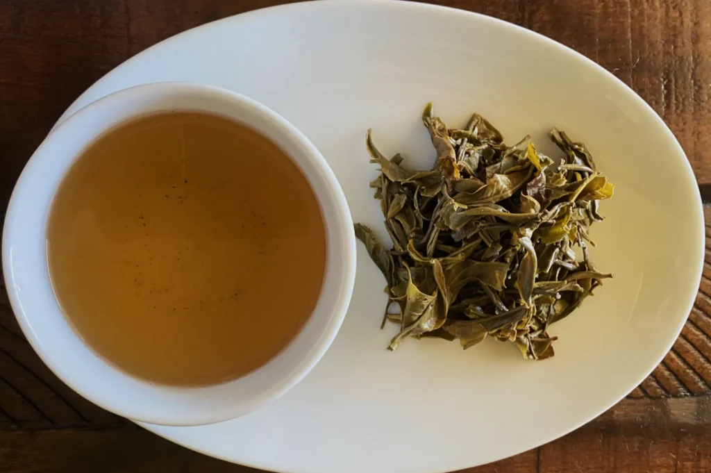 A cup of light amber tea is placed on an oval white plate beside a small pile of brewed green tea leaves, set on a wooden surface, conveying a calming atmosphere.