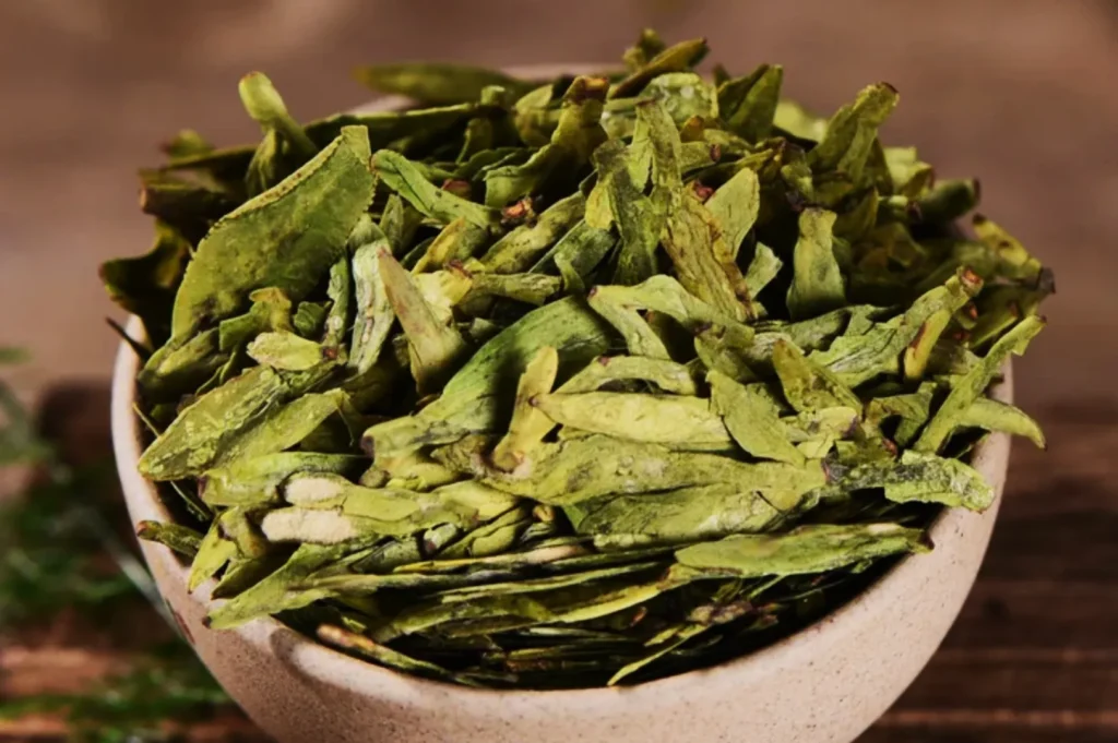 A ceramic bowl filled with vibrant green dried tea leaves, placed on a rustic wooden surface. The leaves are crisp and textured, suggesting freshness.
