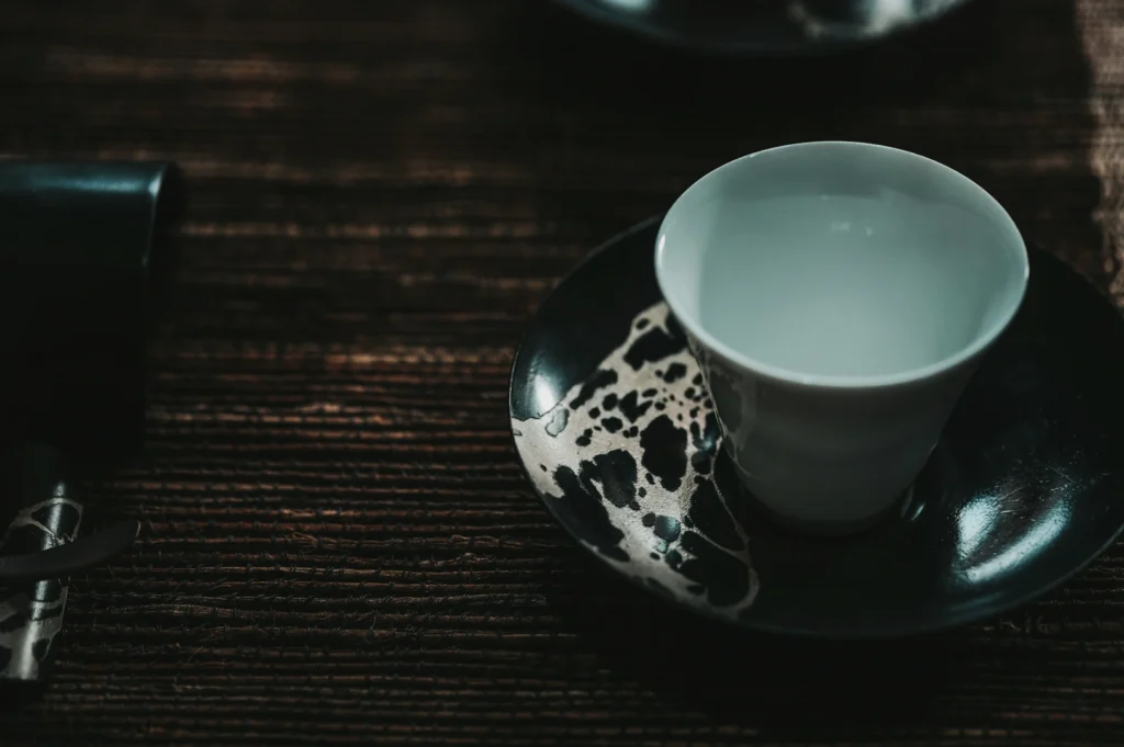 A delicate white cup sits on a black saucer with a unique abstract design, placed on a textured wooden surface, evoking a serene, minimalist ambiance.