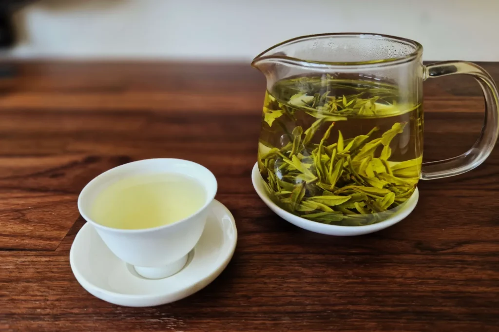 A clear glass teapot with green tea leaves sits on a wooden table, next to a white porcelain cup filled with light green tea, creating a serene, calming atmosphere.