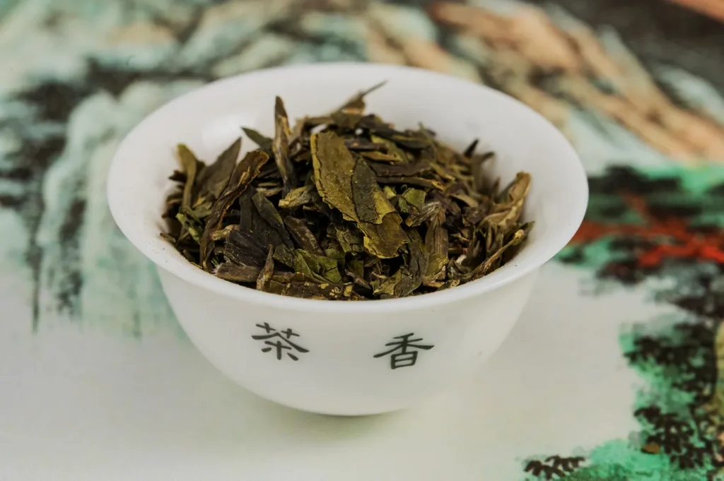A white bowl filled with dried green tea leaves is placed on a colorful, abstract background. The bowl has Chinese characters printed on it.