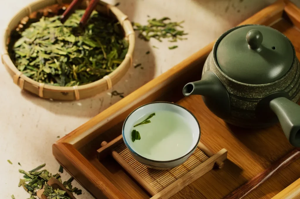 A serene tea setting features a bamboo tray with a green teapot and a small cup of pale green tea accompanied by floating leaves. A basket holds loose tea leaves.