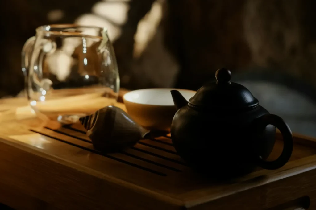 A dark ceramic teapot, a clear glass cup, and a small bowl sit on a wooden tray. Soft light creates a warm, serene atmosphere.