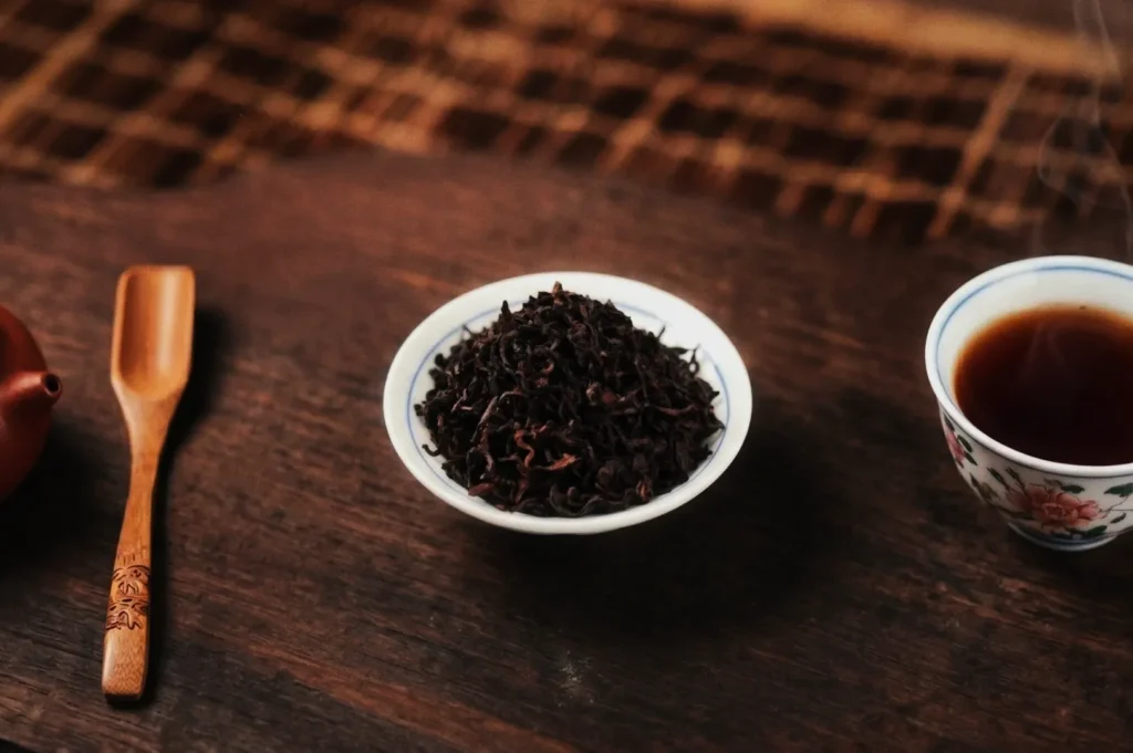 A wooden table displays a small bowl of loose tea leaves, a decorative tea cup with brewed tea, and a carved wooden scoop, conveying a calming atmosphere.