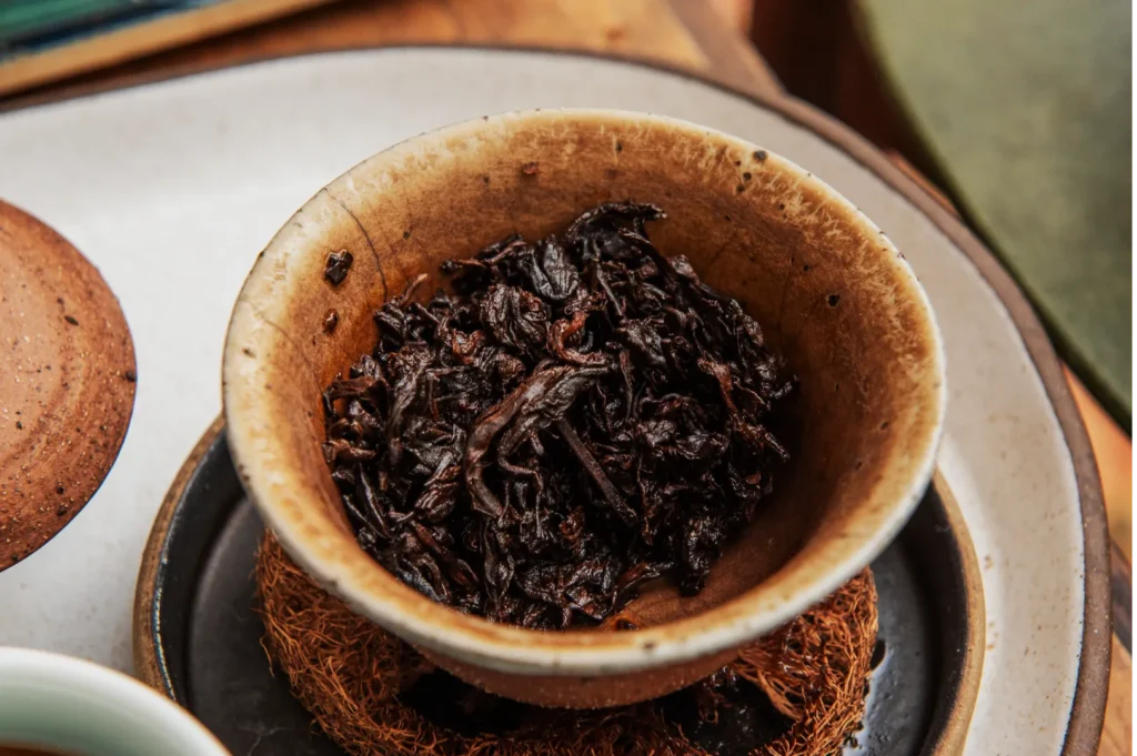 A rustic brown bowl filled with dark, wet tea leaves sits on a textured coaster. The setting conveys warmth and a sense of relaxation.