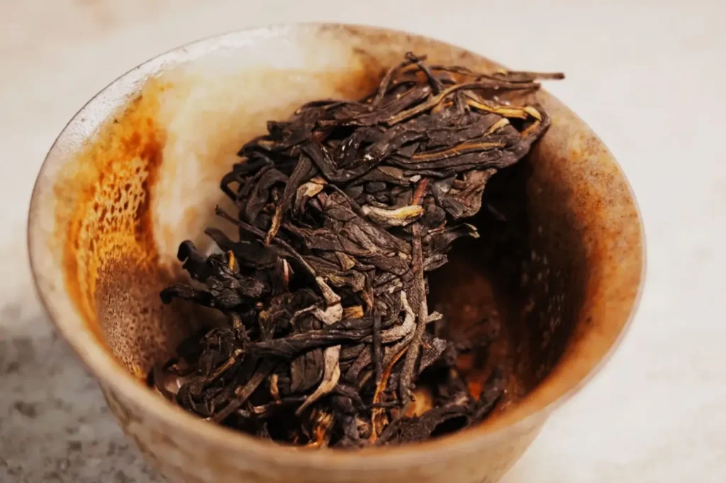 A rustic ceramic bowl holds a pile of dried, dark tea leaves with hints of gold, conveying an earthy and aromatic tone. The background is a soft blur.