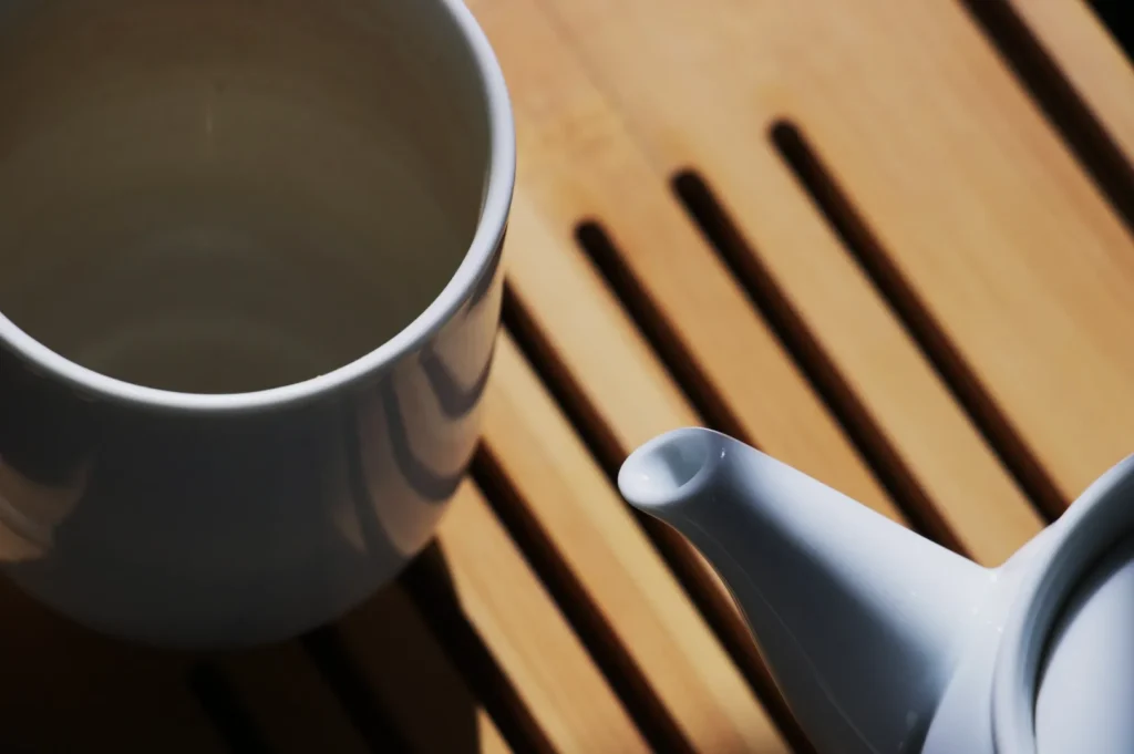 A white teacup and teapot sit on a wooden slatted surface. Sunlight casts soft shadows, creating a warm and inviting atmosphere.