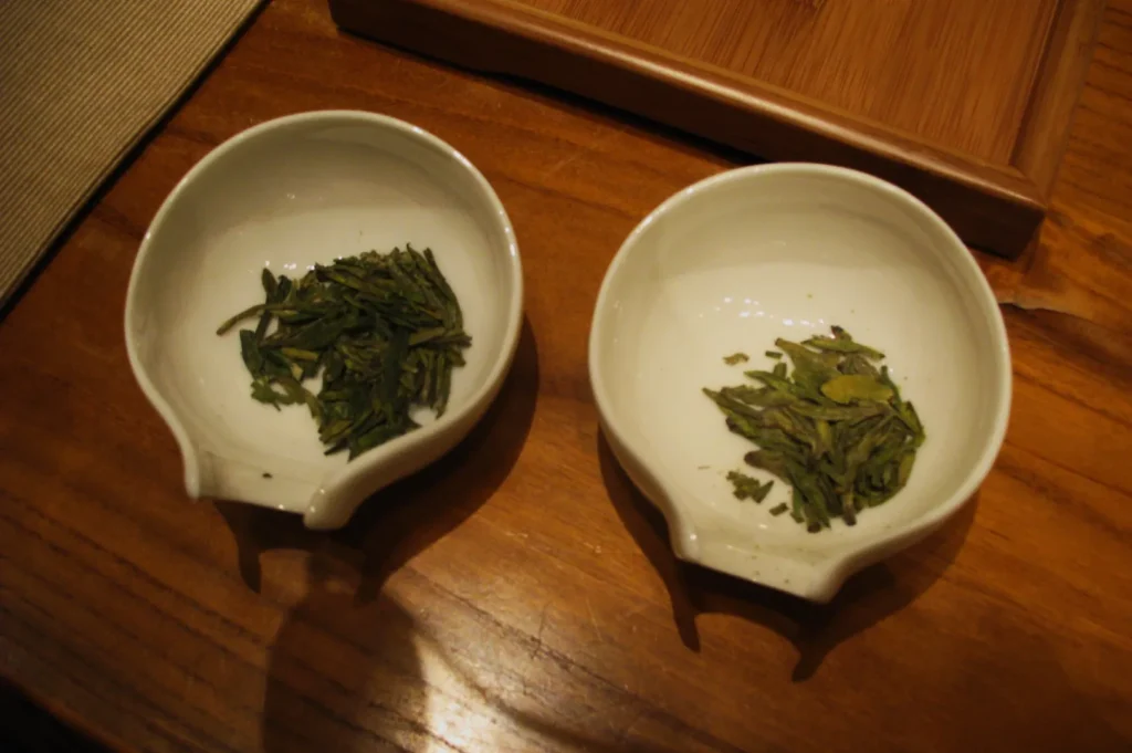 Two small white ceramic bowls on a wooden table contain green tea leaves, slightly varying in size and color, evoking a natural, calming ambiance.