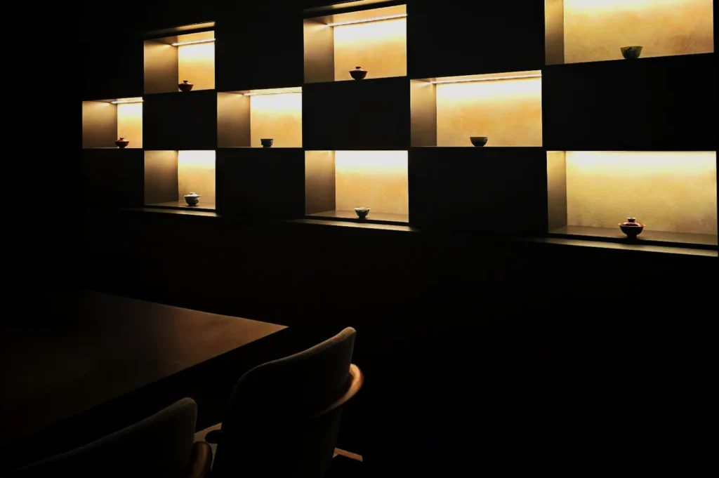 Dimly lit room with a wall of illuminated square shelves, each holding small ceramic bowls. Two empty chairs sit by a table, creating a serene ambiance.