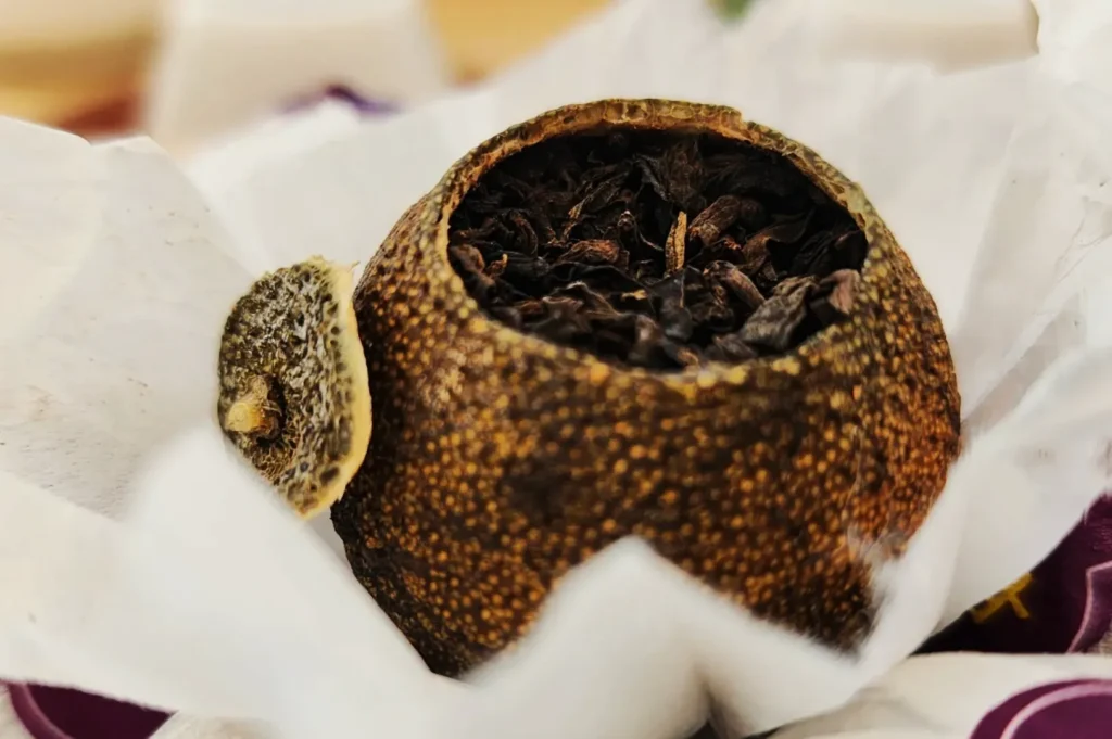 A dried, hollowed citrus peel filled with black tea leaves sits in crinkled white paper, conveying a rustic and aromatic sensory experience.