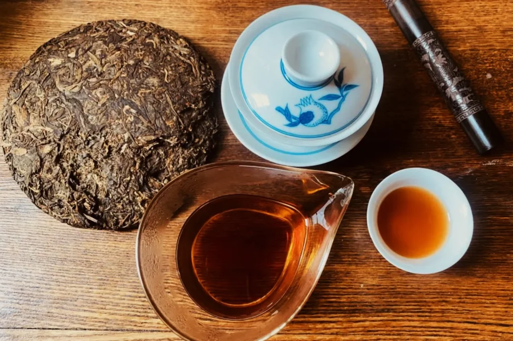 A compressed tea cake, a white teacup with blue designs, a small white saucer, and a clear pitcher of amber tea sit on a wooden table.