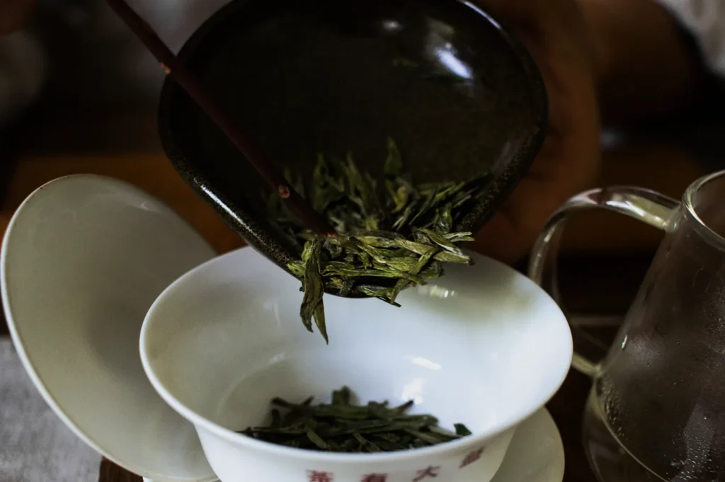 A person uses chopsticks to transfer dried tea leaves from a brown dish into a white teacup with an open lid. A glass pitcher sits nearby.