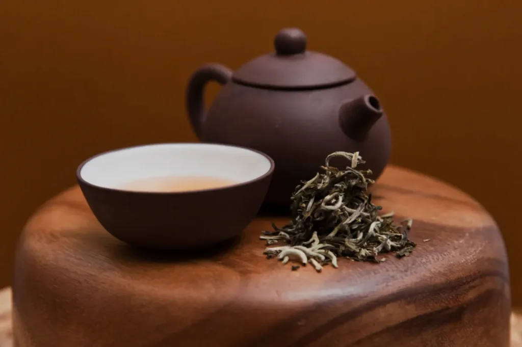 A brown teapot and cup with tea sit on a wooden surface beside a small pile of loose leaf tea. The setting is warm and inviting.