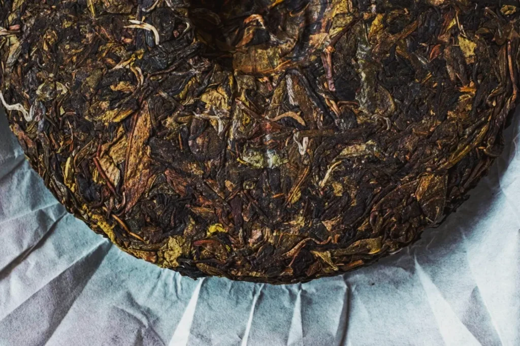 Close-up of a compressed disc of dark, textured tea leaves on crinkled white paper. The leaves are tightly interwoven, displaying a rustic, earthy tone.